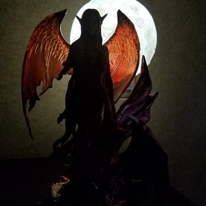 May include: A dark silhouette of a winged figure with a large, glowing moon in the background. The figure has long hair and pointed ears, and is holding a staff. The wings are large and feathered, and are spread out behind the figure.