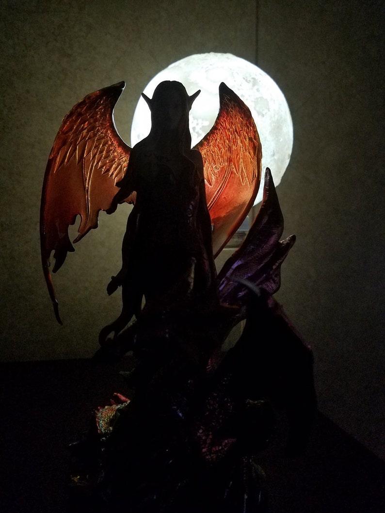May include: A dark silhouette of a winged figure with a large, glowing moon in the background. The figure has long hair and pointed ears, and is holding a staff. The wings are large and feathered, and are spread out behind the figure.