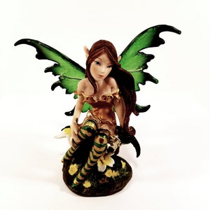 May include: A green-winged fairy figurine with brown hair and a gold and green dress, sitting on a base with flowers.