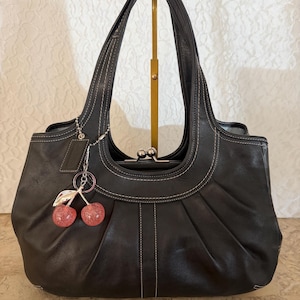 May include: Black leather handbag with white stitching and a silver clasp. The bag has a pleated design and two handles. A cherry charm with pink glitter accents hangs from the handle.