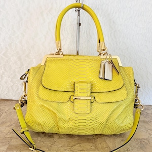 May include: A bright yellow handbag with a snakeskin pattern. The bag has a top handle and a detachable shoulder strap. Gold-colored hardware accents the bag, including the clasp and strap attachments. A small tag hangs from the handle.