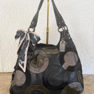 May include: Black leather Coach handbag with a patchwork design of circles and other shapes in shades of gray, black, and olive green. The bag has two black leather handles and a decorative scarf tied to one handle.