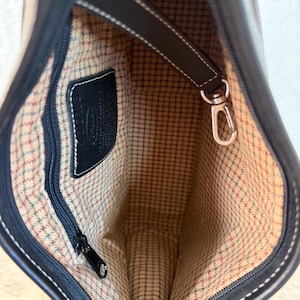 Op de afbeelding: De binnenkant van een donkerbruine leren handtas met een beige, bruine en groene geruite stoffen voering. Het bevat een zwart ritsvak, een zwart leren merklabel en een afneembare bruine leren riem met een zilverkleurige metalen karabijnhaak.