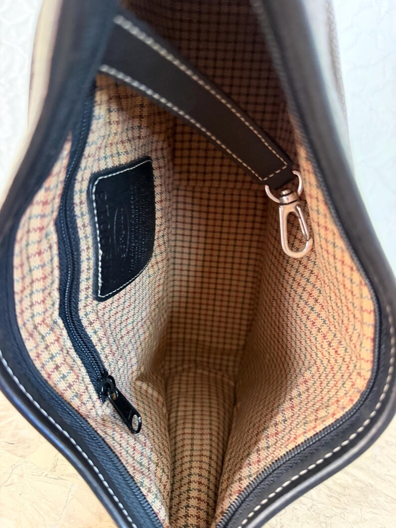 Op de afbeelding: De binnenkant van een donkerbruine leren handtas met een beige, bruine en groene geruite stoffen voering. Het bevat een zwart ritsvak, een zwart leren merklabel en een afneembare bruine leren riem met een zilverkleurige metalen karabijnhaak.