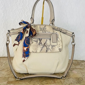 May include: Cream-colored leather handbag with a snakeskin-patterned pocket and a decorative scarf. The bag has a top handle and a detachable shoulder strap. Silver-tone hardware accents the bag's design.