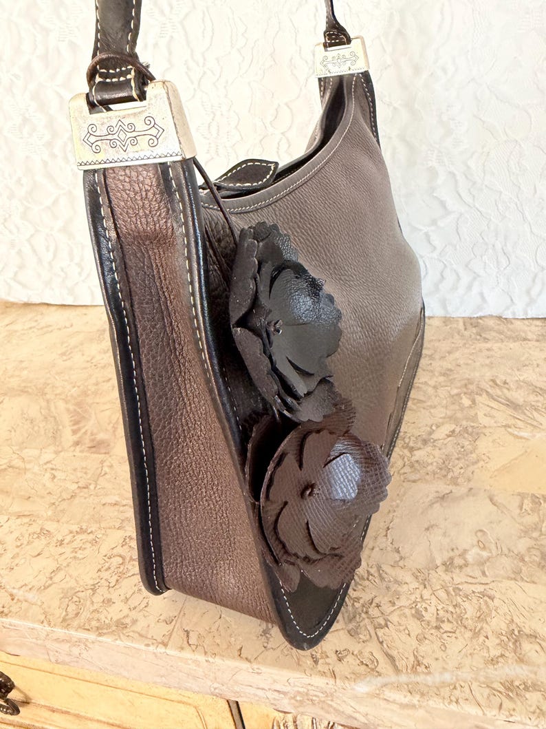 Op de afbeelding: Een donkerbruine leren schoudertas met een getextureerd oppervlak, witte contraststiksels en twee decoratieve donkerbruine leren bloemapplicaties aan de zijkant. De tas is voorzien van zilverkleurige metalen hardware met gegraveerde details.