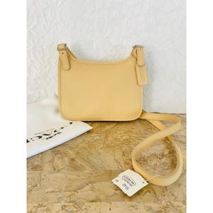 May include: A pale yellow leather crossbody bag with a smooth finish, featuring a long adjustable shoulder strap with silver-toned metal buckles, a rectangular body with rounded corners, and a small rectangular leather hang tag attached to the side.