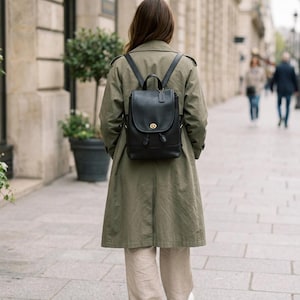 May include: A black leather backpack featuring a top handle, adjustable shoulder straps, and a gold-toned turn-lock closure on the front flap. The bag includes a drawstring opening beneath the flap and a small leather luggage tag attached.