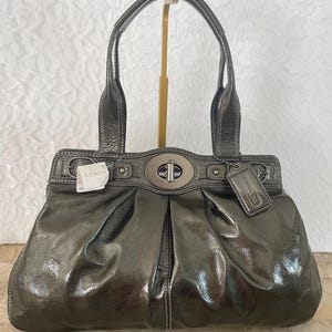 May include: A metallic gray Coach handbag with a top handle and a silver clasp. The bag has a glossy finish and a tag that reads "COACH". The bag is displayed on a gold stand.