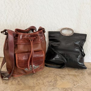 May include: Two leather handbags. One is brown with a drawstring closure and a crocodile-like texture on the front pocket. The other is black with a silver-toned oval handle and a similar texture.