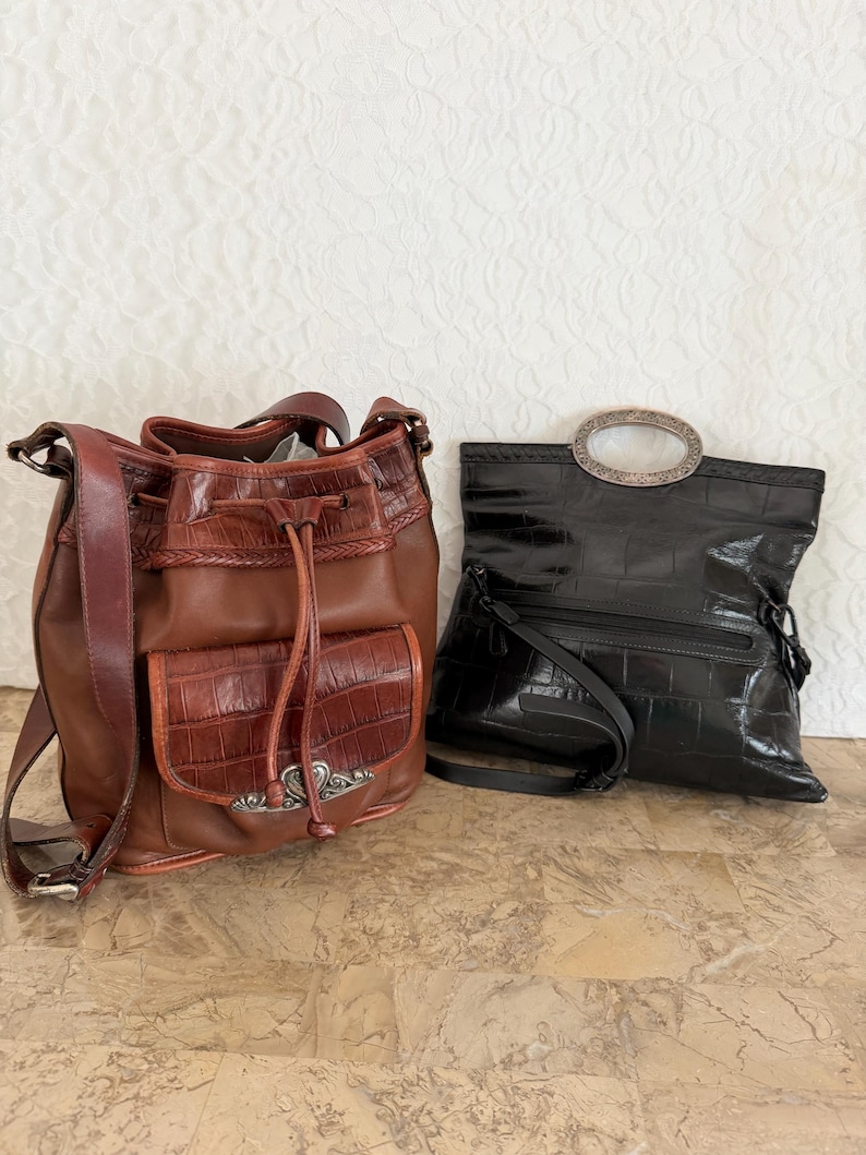 May include: Two leather handbags. One is brown with a drawstring closure and a crocodile-like texture on the front pocket. The other is black with a silver-toned oval handle and a similar texture.