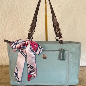 May include: A light blue leather tote bag with brown leather handles, silver-tone hardware, and a front slip pocket. A decorative silk scarf with a nautical rope and wheel print is tied to one handle, and a small leather luggage tag hangs nearby.