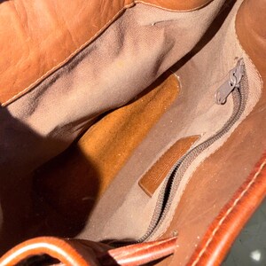 May include: Open view of a brown leather bag's interior. The bag has a drawstring closure and a zippered pocket. The interior lining is a lighter brown fabric. The leather has a rich, warm tone.