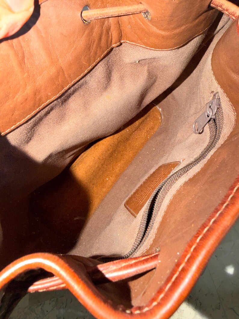 May include: Open view of a brown leather bag's interior. The bag has a drawstring closure and a zippered pocket. The interior lining is a lighter brown fabric. The leather has a rich, warm tone.