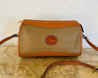 May include: A tan leather crossbody bag with a brown leather strap. The bag has a brown leather flap with a brass buckle and a small brass logo on the front.