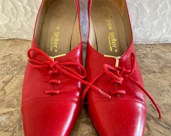 May include: Pair of red leather lace-up shoes. The shoes have a pointed toe and a low heel. The brand name "andrew geller" is visible on the insole. The shoes are a vibrant red color.