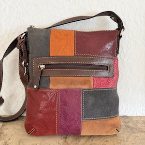 May include: A patchwork crossbody bag with a brown leather strap. The bag features a variety of colors, including red, orange, gray, and purple, in a geometric pattern. The bag has a zippered pocket and a brown leather strap.