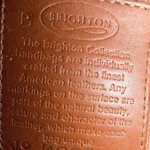 May include: Close-up of a brown leather tag with the embossed word "BRIGHTON" at the top. The tag has text that reads "The Brighton Collection handbags are individually crafted from the finest American leathers." The number "429848" is at the bottom.