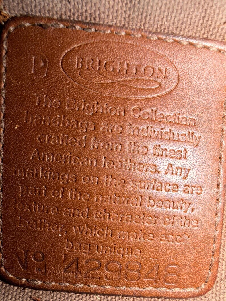 May include: Close-up of a brown leather tag with the embossed word "BRIGHTON" at the top. The tag has text that reads "The Brighton Collection handbags are individually crafted from the finest American leathers." The number "429848" is at the bottom.