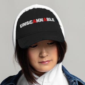 May include: Black baseball cap featuring the word "UNSCANNABLE" embroidered in white and red. The cap has a distressed look and a curved peak. The person in the image is wearing a white hoodie and a denim jacket.