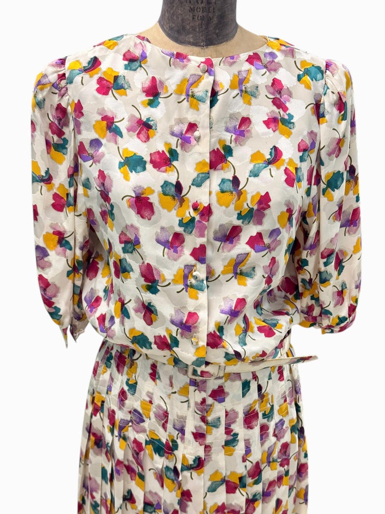 May include: A white dress with a floral print in shades of pink, yellow, green, and purple. The dress has a button-up front and a pleated skirt.