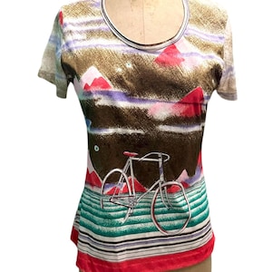 May include: A short-sleeved t-shirt with a bicycle graphic. The shirt features a landscape design with mountains, stripes, and a red bicycle. The bottom hem is red, and the shirt is displayed on a mannequin.