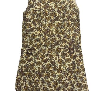 May include: A sleeveless dress featuring a cream background with a dense, multi-colored paisley and floral pattern in shades of brown, dark red, and gold. The garment includes a waistline detail with two small, dark brown rectangular buckles.