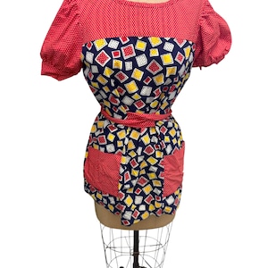 Könnte beinhalten: Vintage-Stil Schürze auf einer Schaufensterpuppe. Das Oberteil ist rot mit weißen Punkten, die Mitte hat ein marineblaues Muster mit gelben und roten Quadraten. Es hat eine rote Taillenbindung und Taschen.