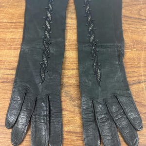 May include: A pair of long black leather gloves featuring a vertical decorative embroidery pattern running down the center of the back of each hand. The gloves have a smooth finish with visible natural grain and stitching details along the fingers.
