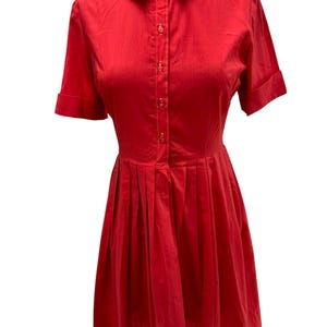 May include: A bright red dress featuring a rounded collar, short sleeves with folded cuffs, and a button-down front placket. The bodice is fitted, transitioning into a pleated skirt that falls from the waistline in a structured silhouette.