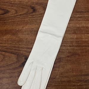 May include: An off-white, elbow-length glove featuring a smooth, matte finish. The glove has four distinct finger sections and a long, straight cuff that extends past the wrist, constructed with visible stitching along the seams of the fingers.