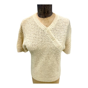 May include: A cream-colored knitted sweater with a v-neck and short sleeves. The sweater has a button closure on the front.