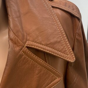 Peut inclure: Gros plan sur une veste en cuir marron avec un grand col et une fermeture &agrave; bouton. La veste a un design unique avec un rabat triangulaire sur le col et des coutures jaunes.