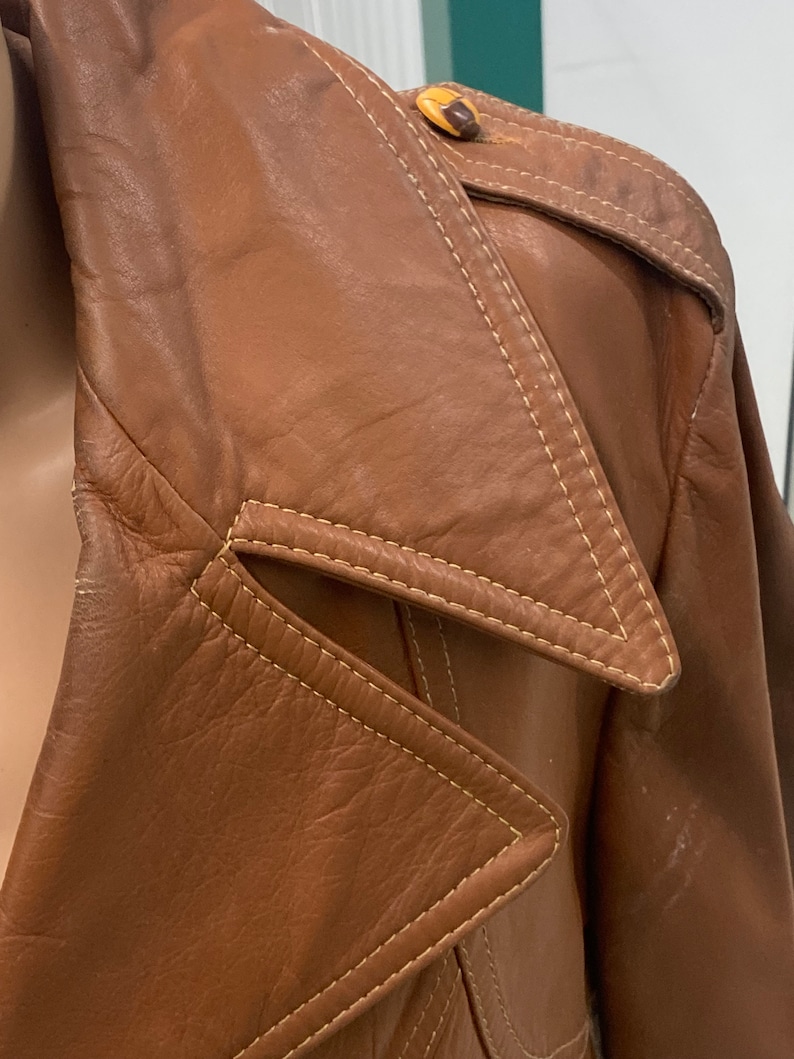 Peut inclure: Gros plan sur une veste en cuir marron avec un grand col et une fermeture &agrave; bouton. La veste a un design unique avec un rabat triangulaire sur le col et des coutures jaunes.