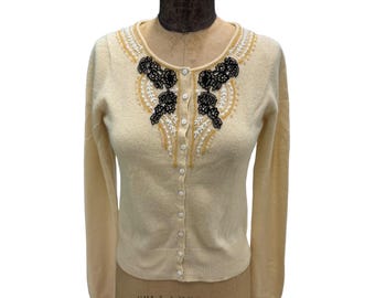 May include: A cream-coloured cardigan with a beaded and embroidered neckline featuring black and gold accents. The cardigan has a button-up front and long sleeves.