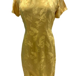 May include: A gold-colored knee-length dress featuring a mandarin collar, short sleeves, and decorative knot closures at the neckline. The fabric displays a tonal jacquard pattern of peacocks and floral motifs throughout the entire garment.