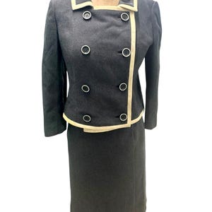 May include: A charcoal grey skirt suit with a double-breasted jacket. The jacket has cream-coloured trim along the collar, lapels, and front edges, with matching buttons. The skirt is a straight, knee-length style. The suit is displayed on a mannequin.