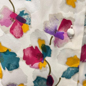 May include: White fabric with a floral pattern. The flowers are painted in shades of pink, purple, yellow, teal, and green. The fabric has a textured surface.