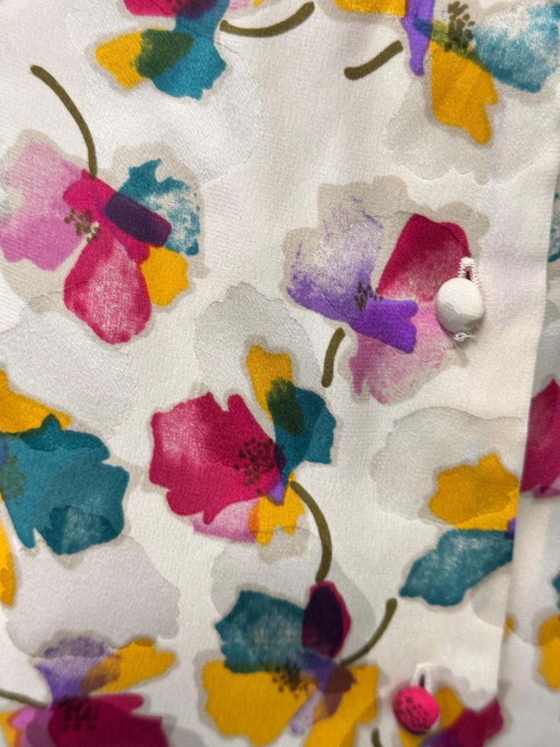 May include: White fabric with a floral pattern. The flowers are painted in shades of pink, purple, yellow, teal, and green. The fabric has a textured surface.