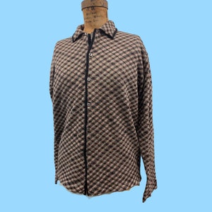 May include: A long-sleeved button-down shirt with a diamond pattern in shades of brown, black, and tan. The shirt has a black collar and trim, and is displayed on a mannequin. The background is a solid light blue.
