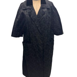 May include: A black, knee-length coat with short sleeves and a velvet collar. The coat has a textured pattern and two front pockets. It is displayed on a mannequin against a white background.