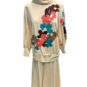 May include: A vintage cream-colored two-piece outfit featuring a long-sleeved top and a skirt. The top has a cowl neck and is adorned with large, colorful floral appliqués in shades of brown, coral, and teal. The skirt is a simple, knee-length design.