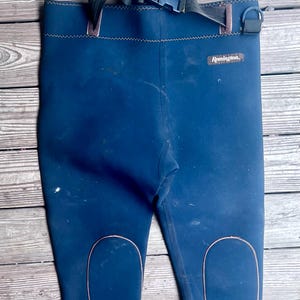 May include: Blue waders with reinforced knee patches and a black adjustable belt. The waders have the brand name "Remington" printed on them. The waders are designed for fishing or water activities.