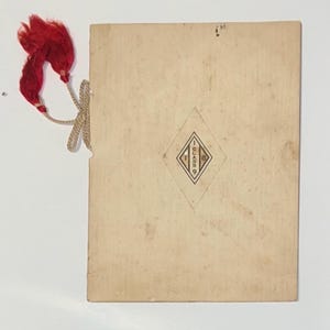 Könnte beinhalten: Eine rechteckige Vintage-Karte mit einem rautenförmigen Emblem und dem Text "GLASS 9". Eine rote Quaste ist mit einer beigen Schnur an der Karte befestigt. Die Karte hat ein abgenutztes, gealtertes Aussehen.