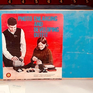 May include: A vintage blue box with the words "PHOTO ENLARGING AND DEVELOPING OUTFIT" in white and red. The box features a retro image of a man and woman working with photography equipment. The background is a dark red.