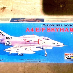 以下が含まれることがあります： マクドネル・ダグラス A4 E/F スカイホークのモデルキットが入った青い長方形の段ボール箱。箱には、A4 E/F SKYHAWK、USS BON HOMME RICHARD、311という文字が書かれた白い海軍ジェット機のグラフィック、Minicraftのロゴ、1/72スケールの表記が印刷されています。