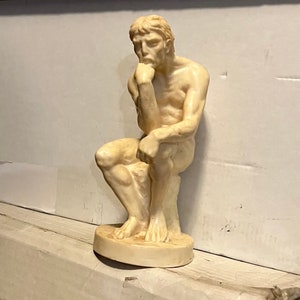 May include: A white marble sculpture of a man sitting with his chin resting on his hand, in a contemplative pose.