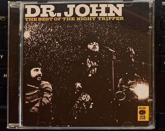 Puede incluir: Carcasa de CD del álbum "The Best of the Night Tripper" de Dr. John. La portada, en tonos marrones y negros, muestra una foto de la banda y el título en blanco. Un adhesivo "Deluxe 2CD" se encuentra en la esquina inferior derecha.