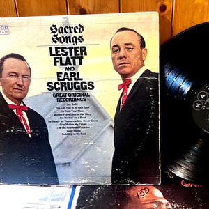 May include: A vinyl record and its square cardboard sleeve featuring Lester Flatt and Earl Scruggs. The cover displays the title Sacred Songs, the artists' names, and a tracklist. The black vinyl record has a circular Harmony label in the center.