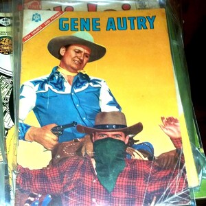 Op de afbeelding: Vintage stripboekcover met Gene Autry in cowboy-outfit, met een pistool. De cover heeft een gele achtergrond met de titel "GENE AUTRY" in blauw. Een andere cowboy draagt een rood geruit overhemd en een groene bandana.